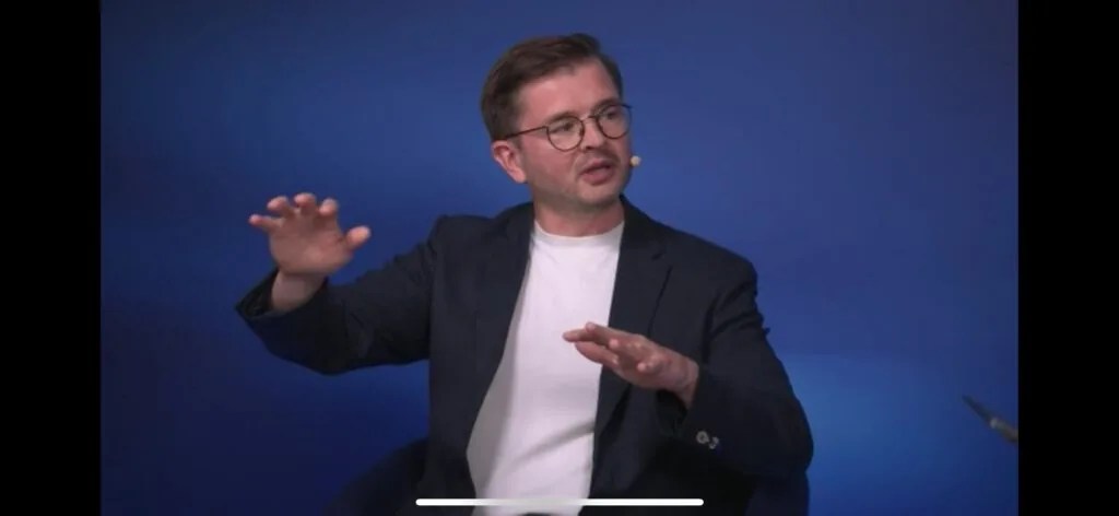 Kevin Cremanns gesturing while explaining a point in front of a blue studio background