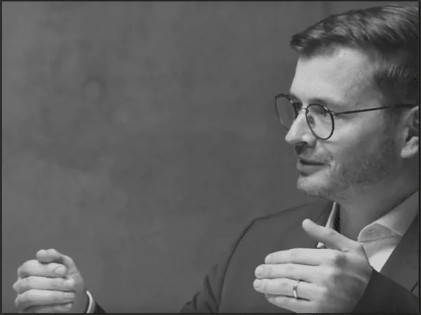 Black-and-white still of a presenter with glasses gesturing while speaking during the CADFEM Blueprint video series