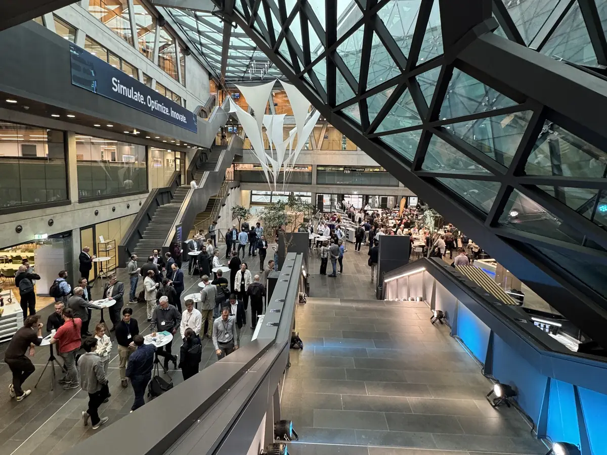 View from the upper floor into the crowded atrium under the glass roof, with a banner reading Simulate. Optimize. Innovate.