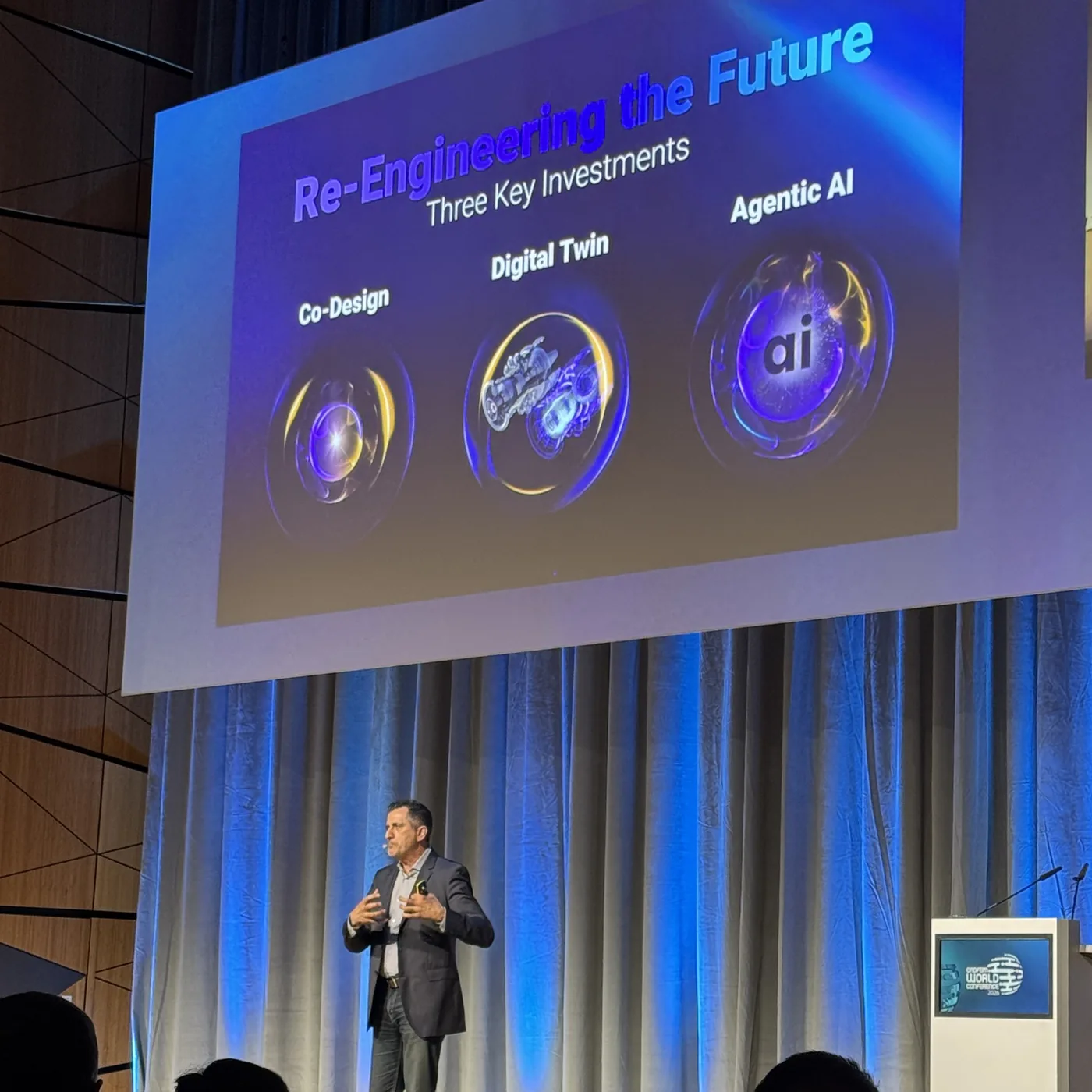 A speaker on stage under a slide titled Re-Engineering the Future, Three Key Investments, naming Co-Design, Digital Twin and Agentic AI.