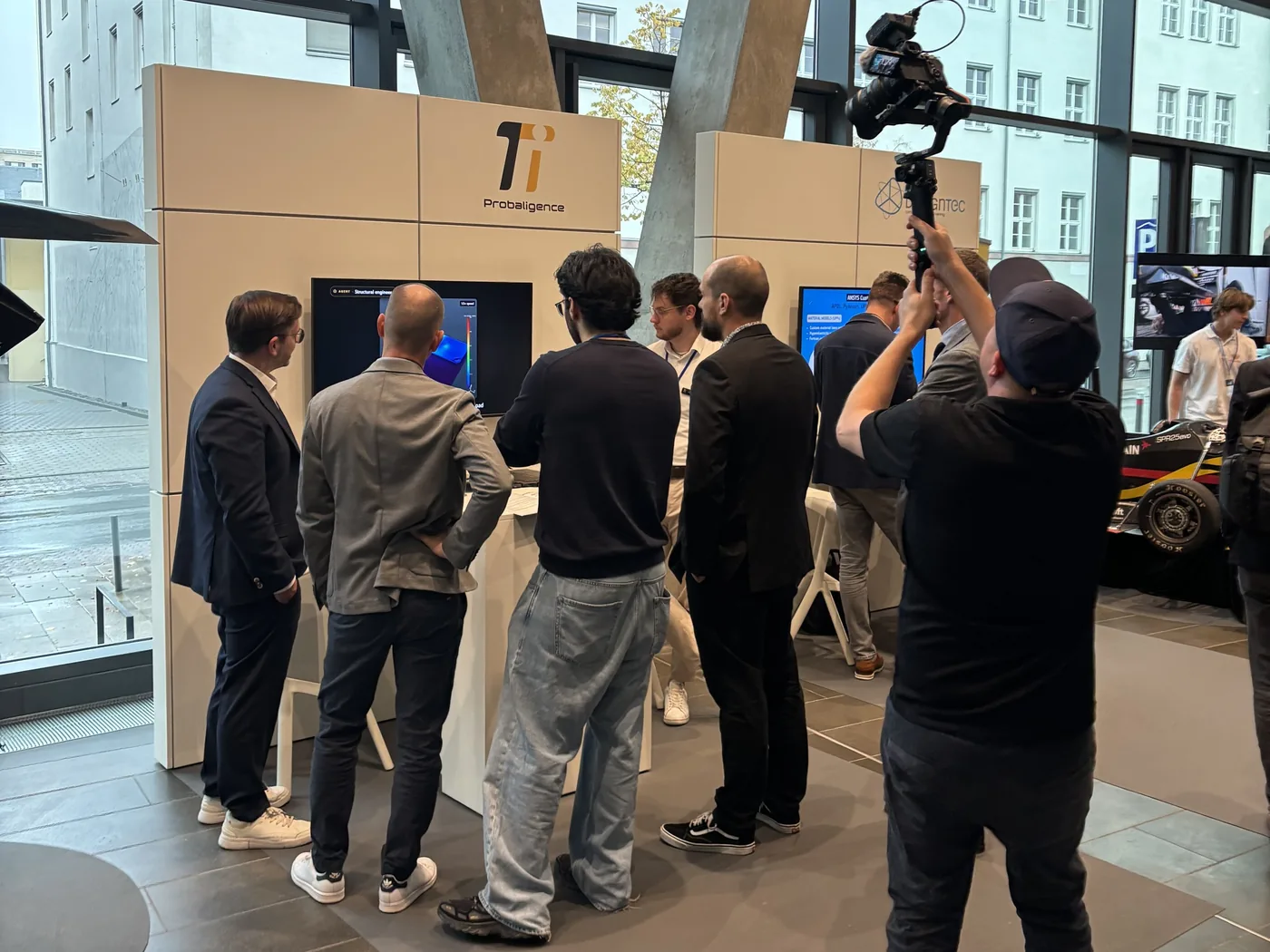 Visitors at the PI Probaligence booth watching a demo on the screen below the Probaligence logo, while a camera operator films the scene.