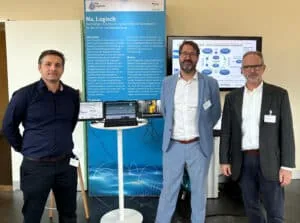 Three consortium members standing at the Na, Logisch exhibition stand in front of the project poster and a laptop demo