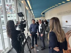 Two exhibitors being interviewed on camera by a film crew in a window-lined hall at the Berlin congress center