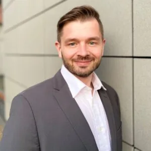 Portrait of Dr. Cremanns in a grey suit jacket and white shirt, standing in front of a tiled building facade