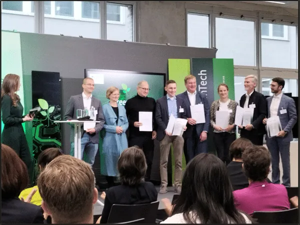 Nine project representatives standing on a stage at the Green Tech kick-off event in Berlin, several holding certificate folders, with an audience in the foreground.