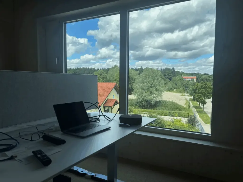 Height-adjustable desk with a laptop in front of large windows overlooking trees and the surroundings of Grafing
